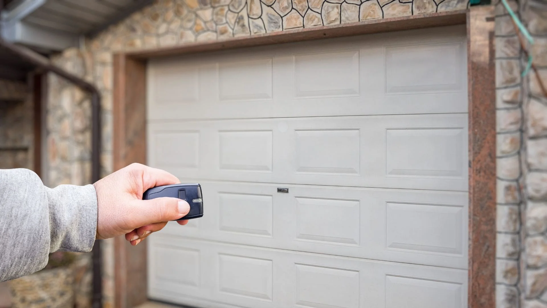 Hand holding garage door remote control in front of white raised-panel garage door with stone surround